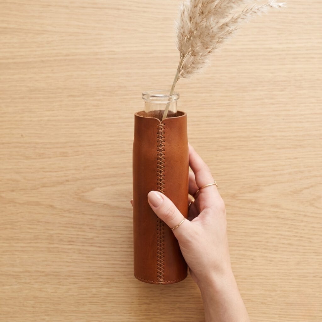 A manicured hand holding a handcrafted saddle-leather solitary sleeve vase, created from a narrow upcycled glass bottle.