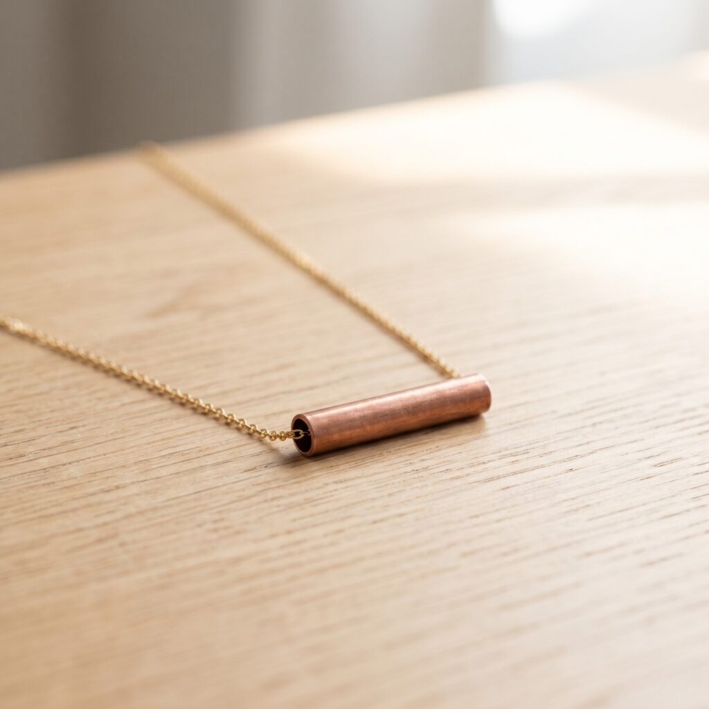 A perfectly square photo showing a sleek raw copper tube pendant sliding on a delicate gold chain, resting on a light oak wood table surface under soft diffused summer sunlight. Industrial aesthetic.