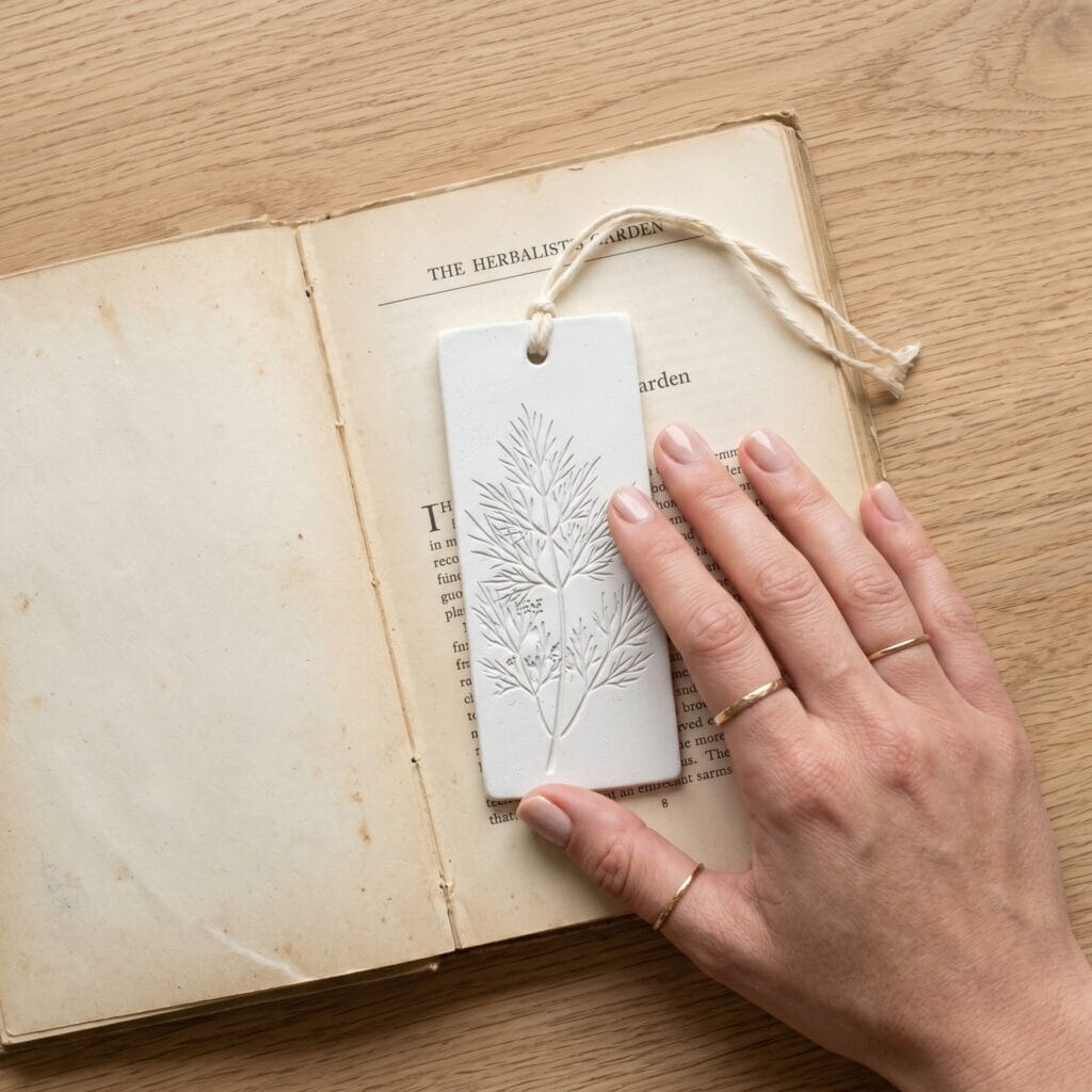 A manicured hand placing an ultra-thin, flat white plaster bookmark with delicate bas-relief dill frond imprints onto an open vintage book.