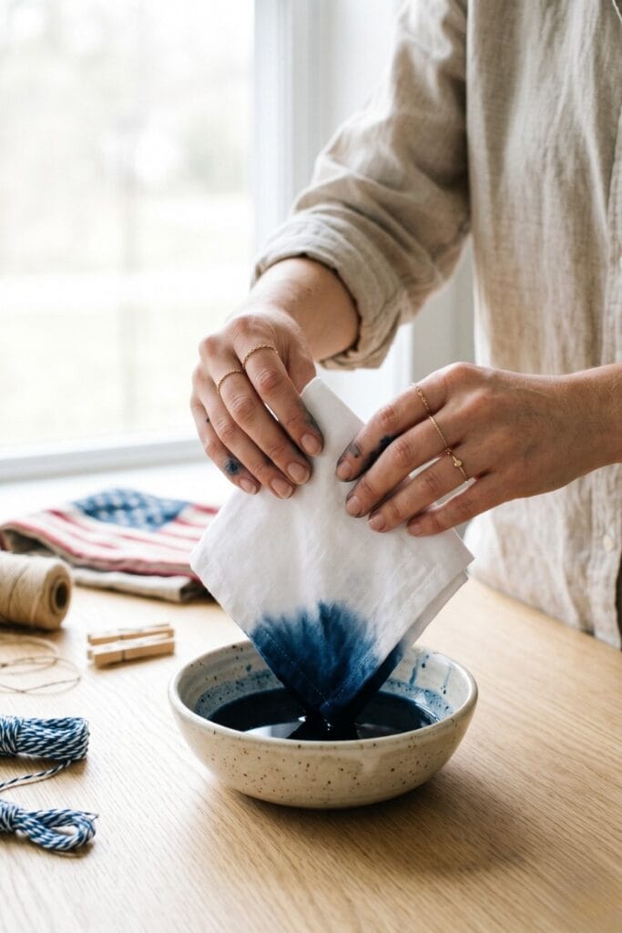 Female artisan hands dip-dying white linen into indigo blue for 4th of July crafts.