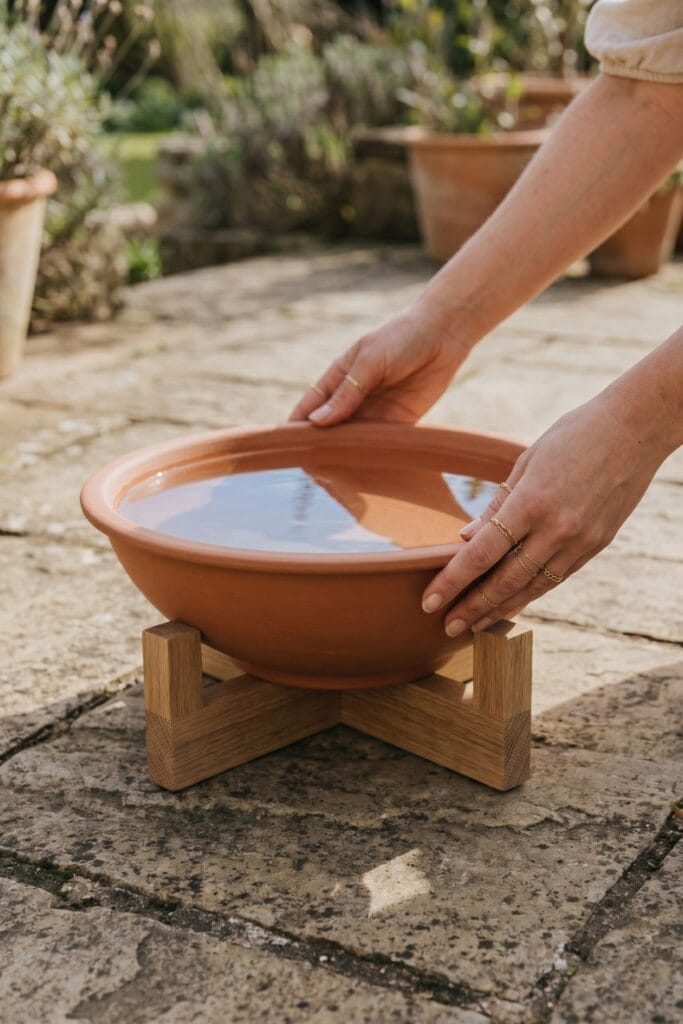 Female artisan hands adjusting a finished minimalist water station made of a wide matte terracotta bowl on a low wooden base filled with water.