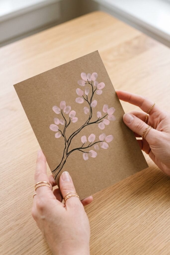 Female artisan hands holding a completed spring daycare crafts artwork on brown kraft paper, displaying a black branch with soft pink fingerprint cherry blossoms.