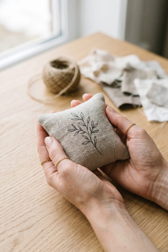 Close-up of female artisan hands holding a single completed small square raw linen pouch stuffed with fiber fill and featuring a delicate single-line black ink botanical pattern.