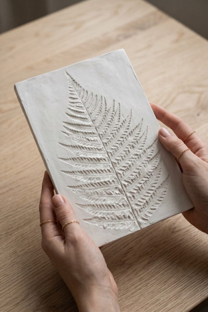Female artisan hands holding a completed rectangular unpainted white plaster cast featuring a detailed imprint of a large spring leaf.