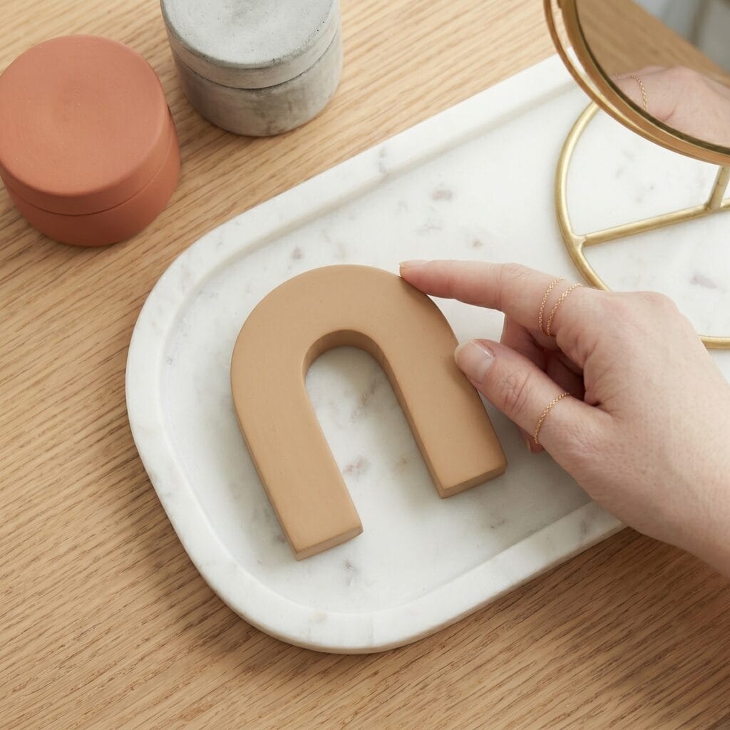 A hand resting on the top curve of a finished, smooth tan polymer clay monolithic arch sculpture displayed on a white marble tray.