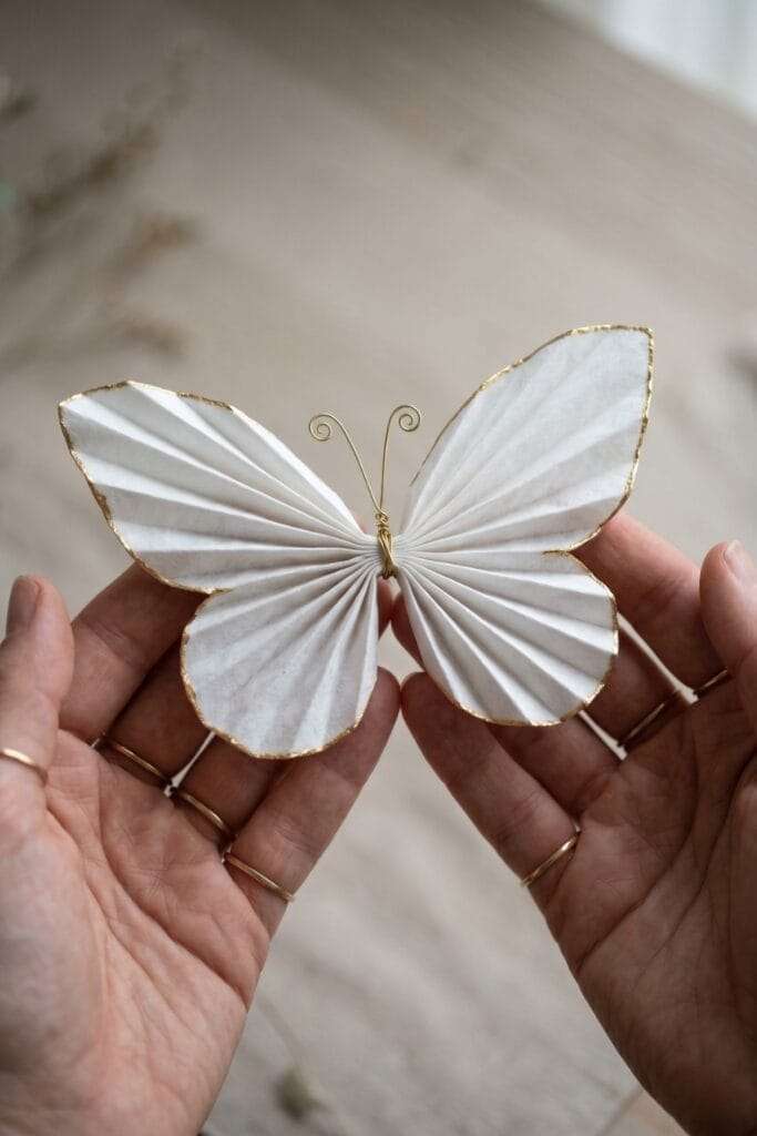 Female artisan hands with a single fingertip with gold chain rings gently rubbing metallic gold wax along the edge of a white coffee filter wing.