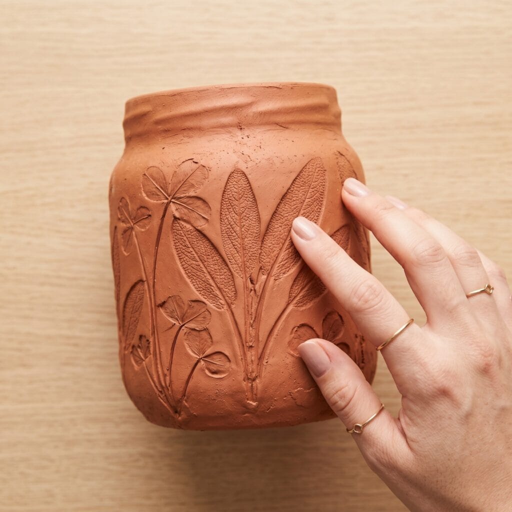 A close-up of a manicured hand tracing tactile bas-relief botanical leaf impressions on a terracotta pot made from an upcycled glass jar.