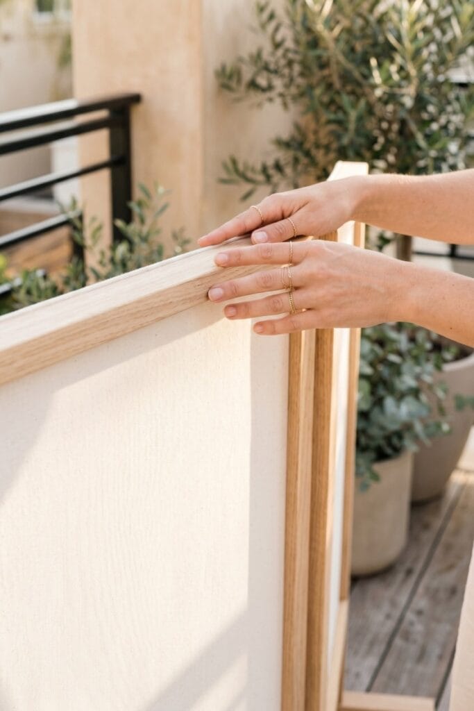 Female artisan hands touching the edge of a finished minimalist outdoor privacy screen made of a raw wood frame and taut natural white canvas.