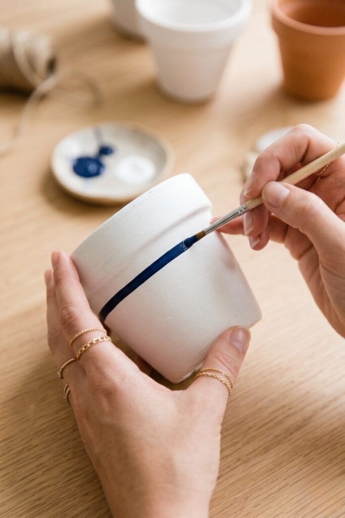 Female artisan hands painting a navy blue stripe on a white terracotta pot for 4th of July crafts.