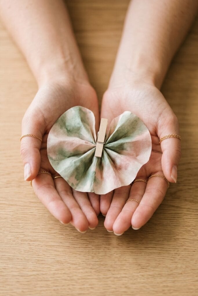 Female artisan hands holding a completed spring daycare crafts project showing a coffee filter painted with soft watercolor pastels and pinched with a wooden clothespin.