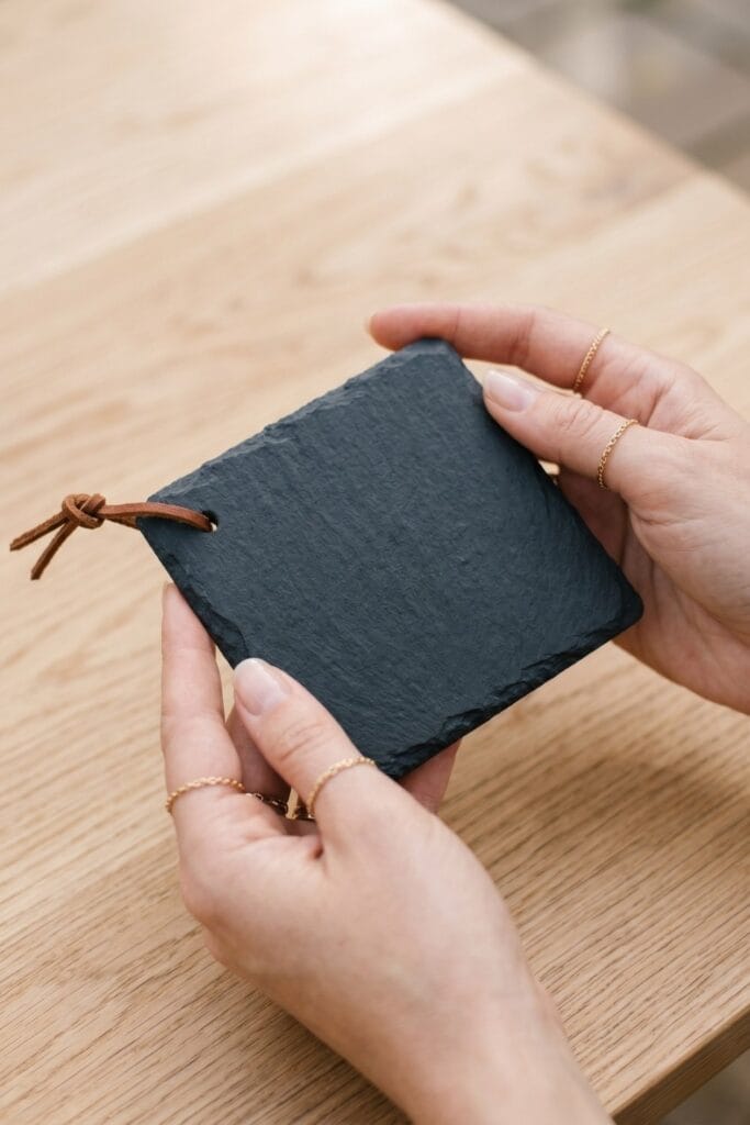 Female artisan hands holding a completed natural dark slate outdoor coaster featuring a thin leather cord tie through a corner hole.