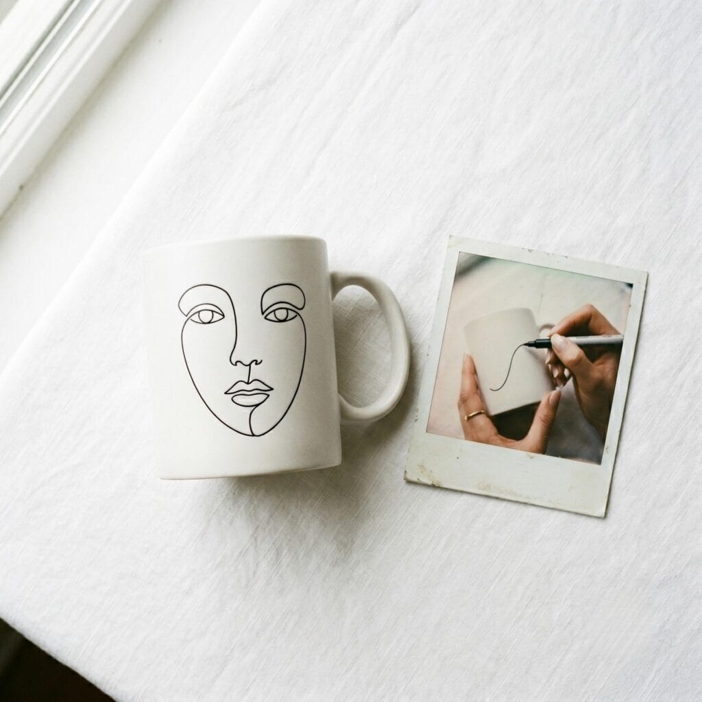 A white ceramic mug featuring a minimalist black abstract line art face on a white table next to a polaroid showing a hand drawing the lines.