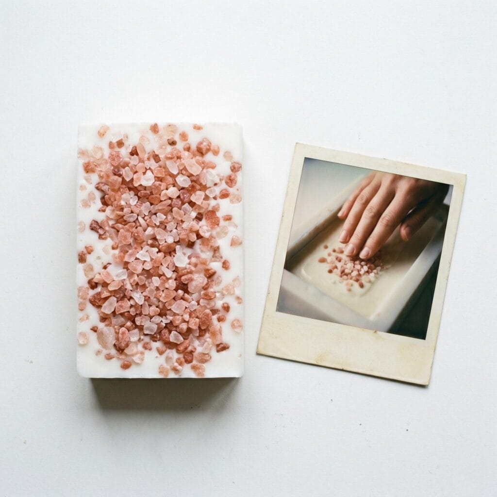 A white soap bar with pink salt crystals on top on a white table next to a polaroid showing a hand pressing crystals onto a cooling soap surface.