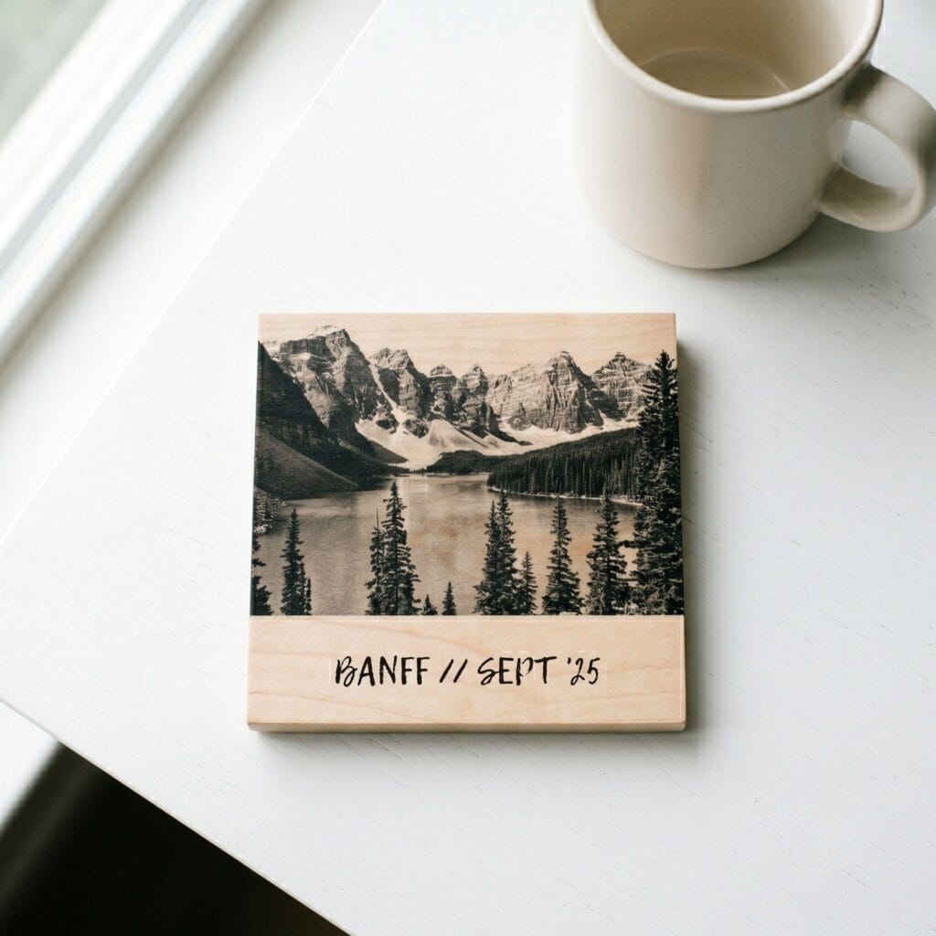 A square light maple wood coaster on a clean white desk, featuring a transferred black-and-white mountain landscape photo and handwritten text reading BANFF SEPT 26.