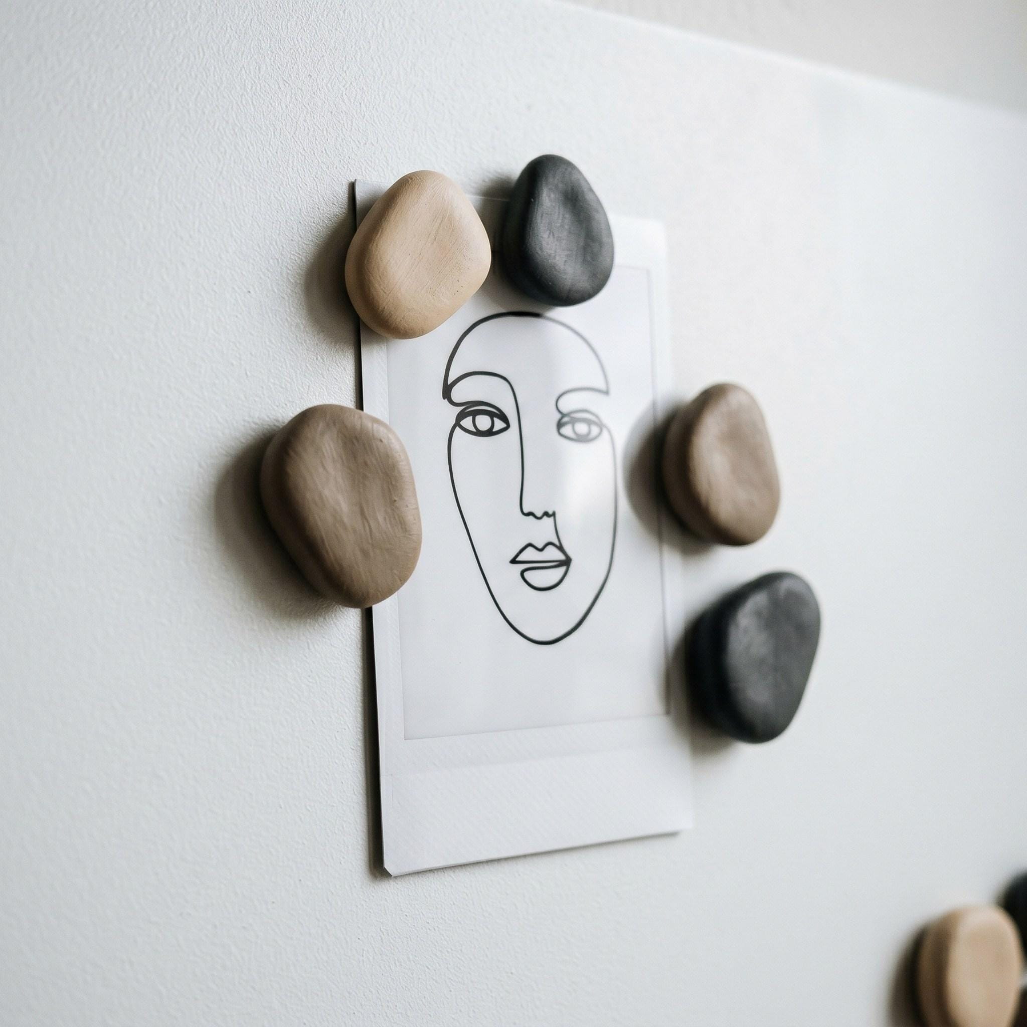 Freehand pebble-shaped clay magnets painted in charcoal and beige tones holding a polaroid photo on a metal board.