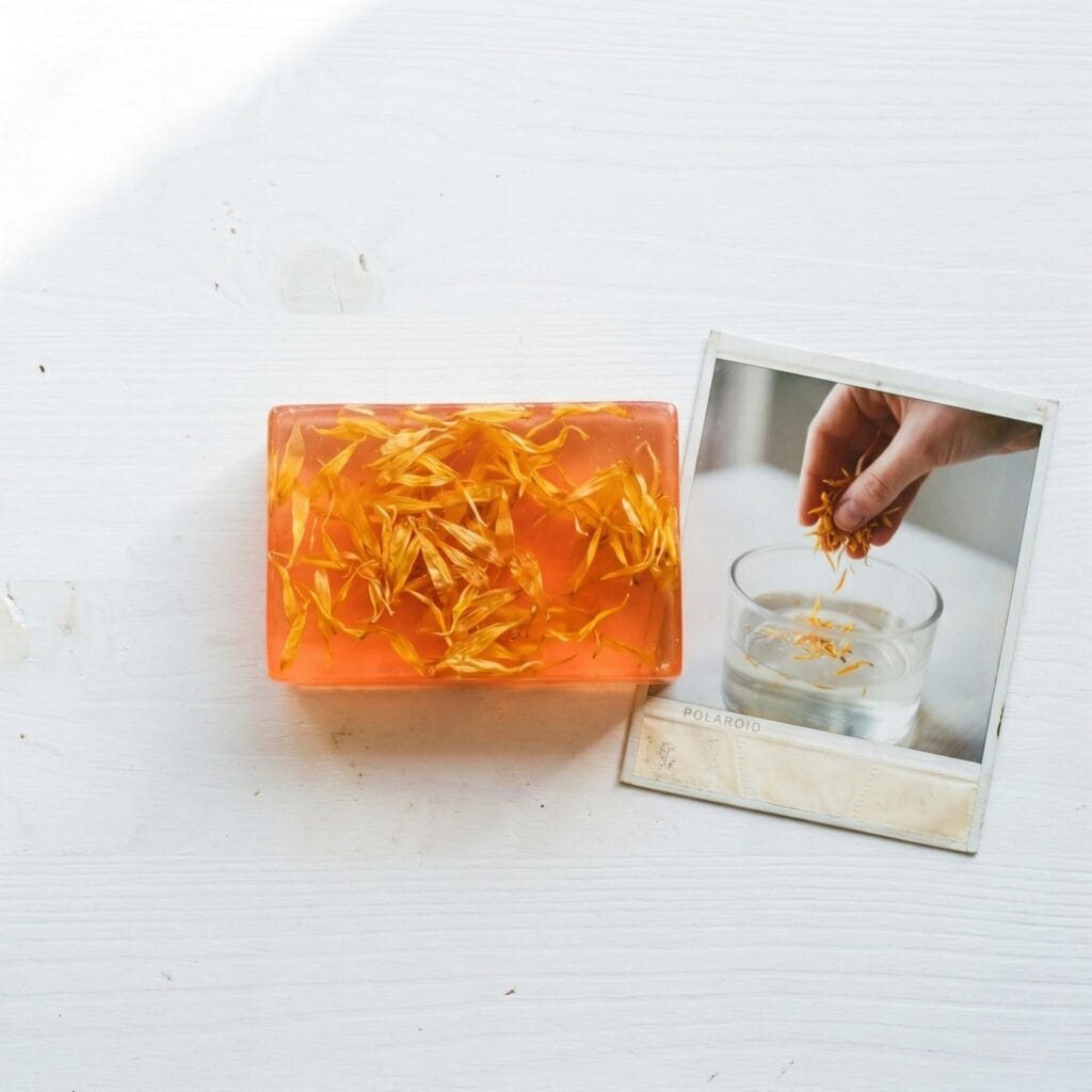 A translucent orange glycerin soap bar with visible calendula petals inside on a white table next to a polaroid showing a hand dropping petals into liquid soap.