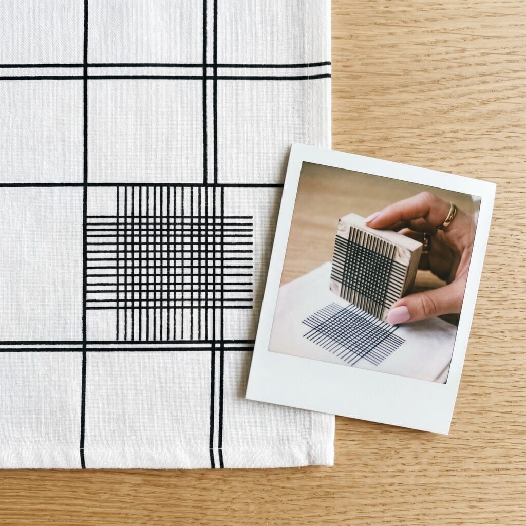A minimalist white cotton napkin showcasing a clean intersecting black monoline grid pattern next to a polaroid photo capturing a hand holding a square stamp with physically raised lines on its base