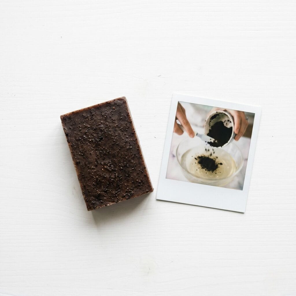 A dark brown coffee soap bar on a white table next to a polaroid showing a hand scraping used coffee grounds into a clear soap base.