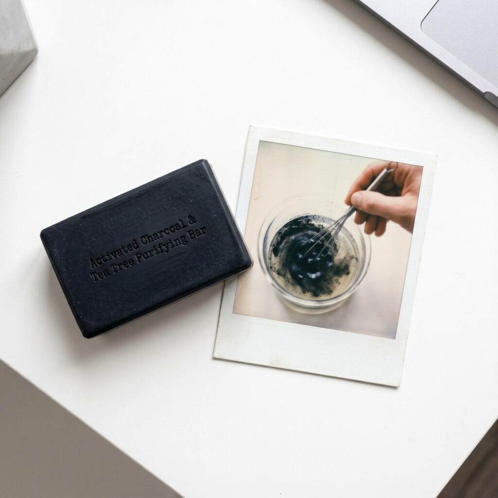 A matte deep black charcoal soap bar on a white table next to a polaroid showing a hand whisking black charcoal powder into a melted base.