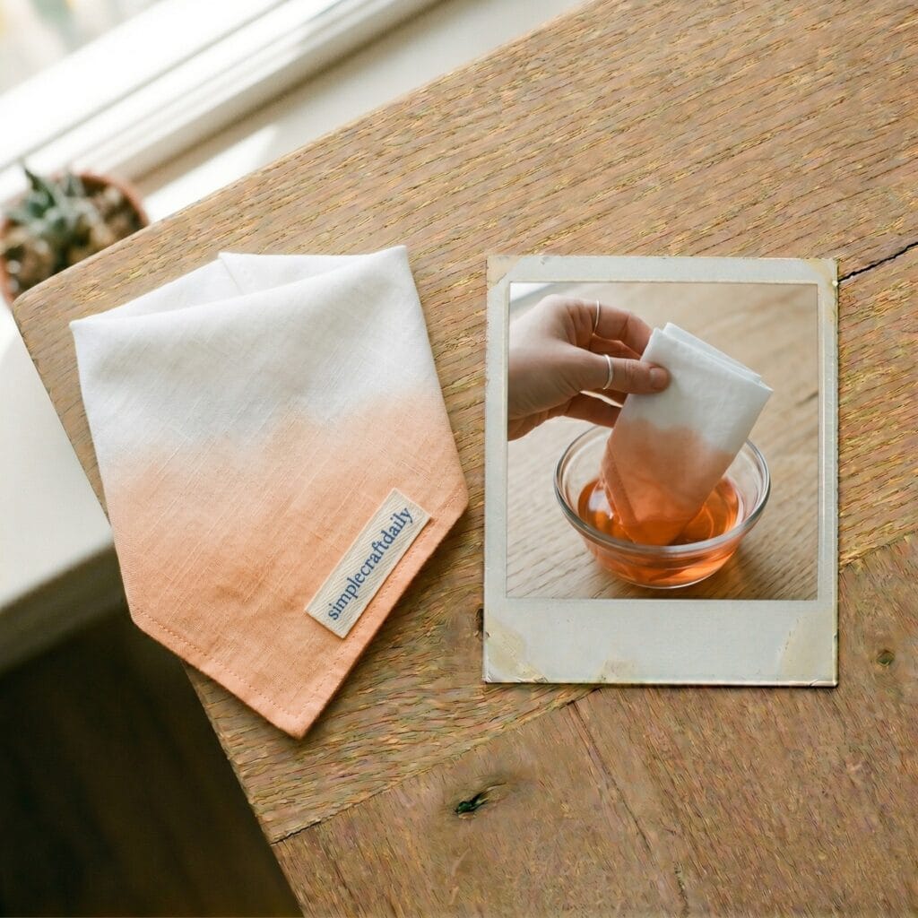 A peach ombre gradient dog bandana on a light oak table next to a polaroid photo showing a single hand dipping white cotton fabric into a bowl of natural organic dye.