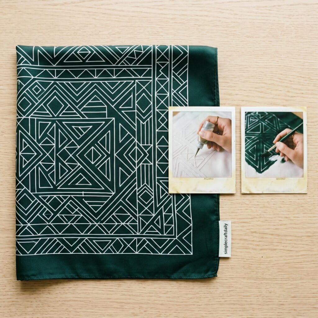 A dark green silk bandana with crisp white geometric lines on a wooden table, next to two vintage polaroid photos: one showing a hand drawing resist paste lines, and the second showing a hand painting green dye over the pattern.