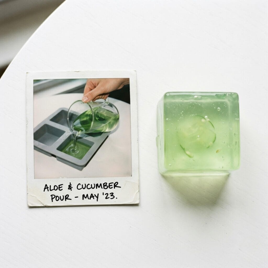 A translucent light green aloe vera soap cube on a white table next to a polaroid showing a hand pouring green liquid into a square mold.