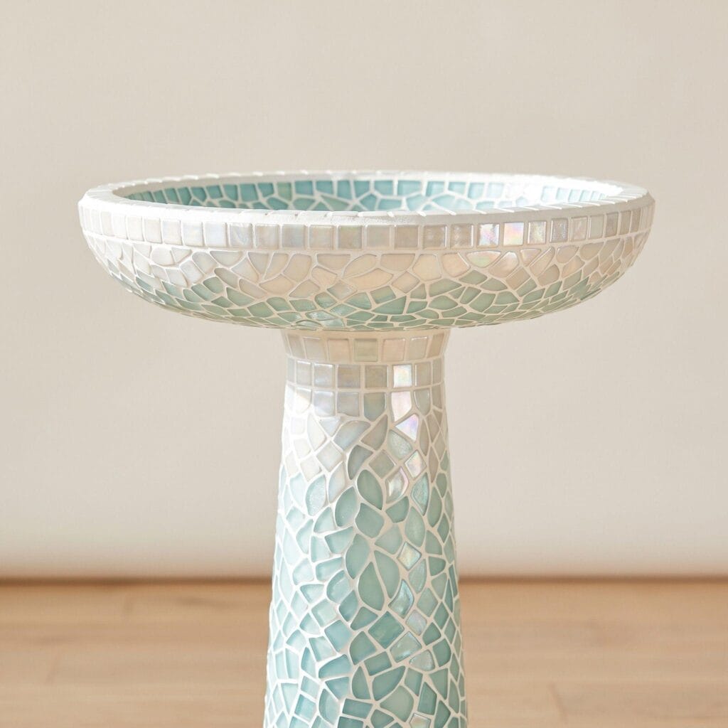 An outdoor concrete birdbath pedestal and rim decorated with detailed glass mosaic tiles.