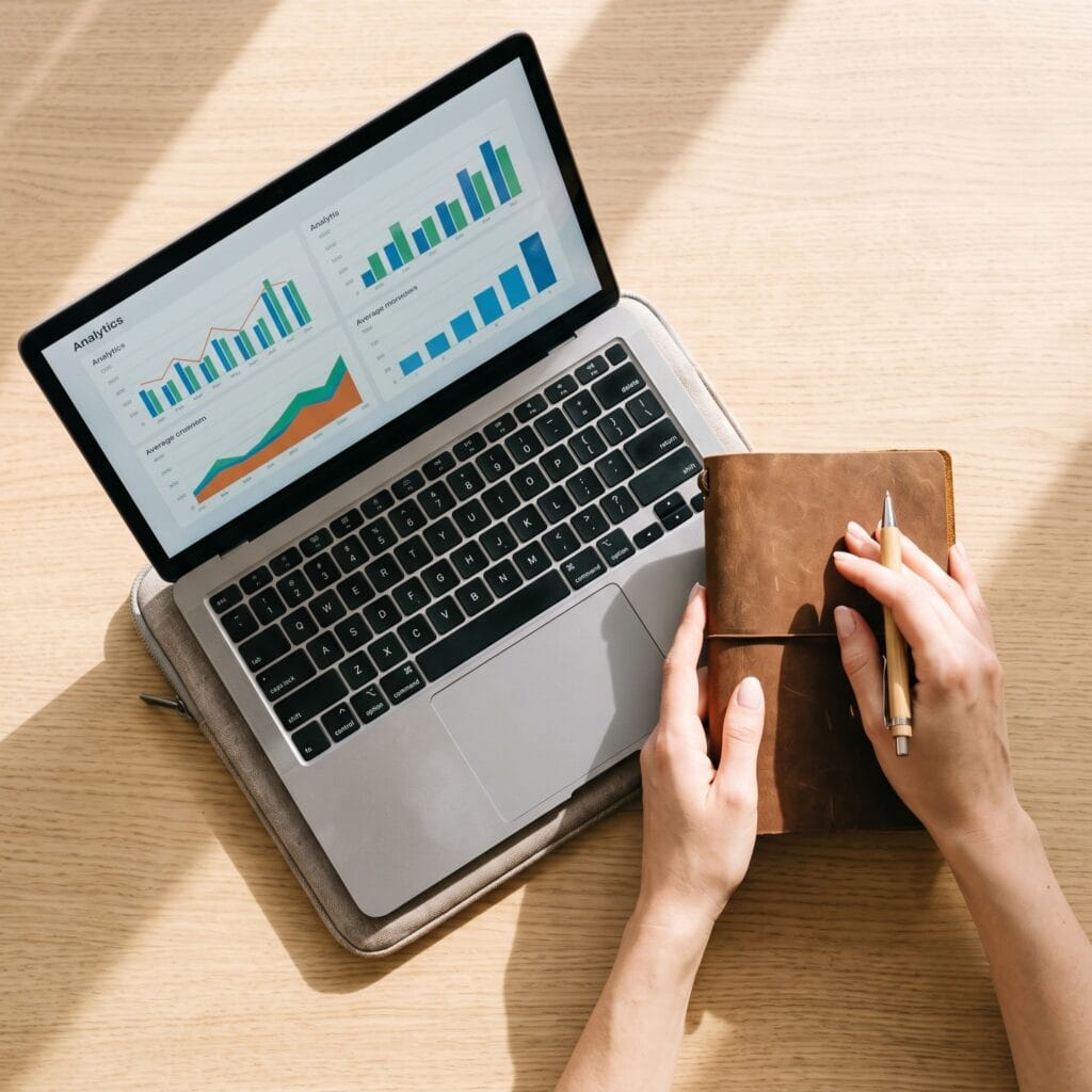 Top-down flat lay view of a laptop displaying shop analytics graphs next to a handmade leather notebook on a wooden desk.