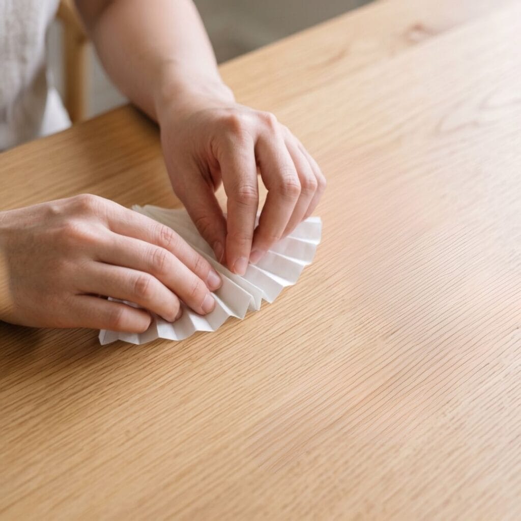 Female artisan hands actively accordion-folding a white paper coffee filter into narrow, even pleats on a light oak wood table, demonstrating the base technique for a DIY coffee filter butterfly.