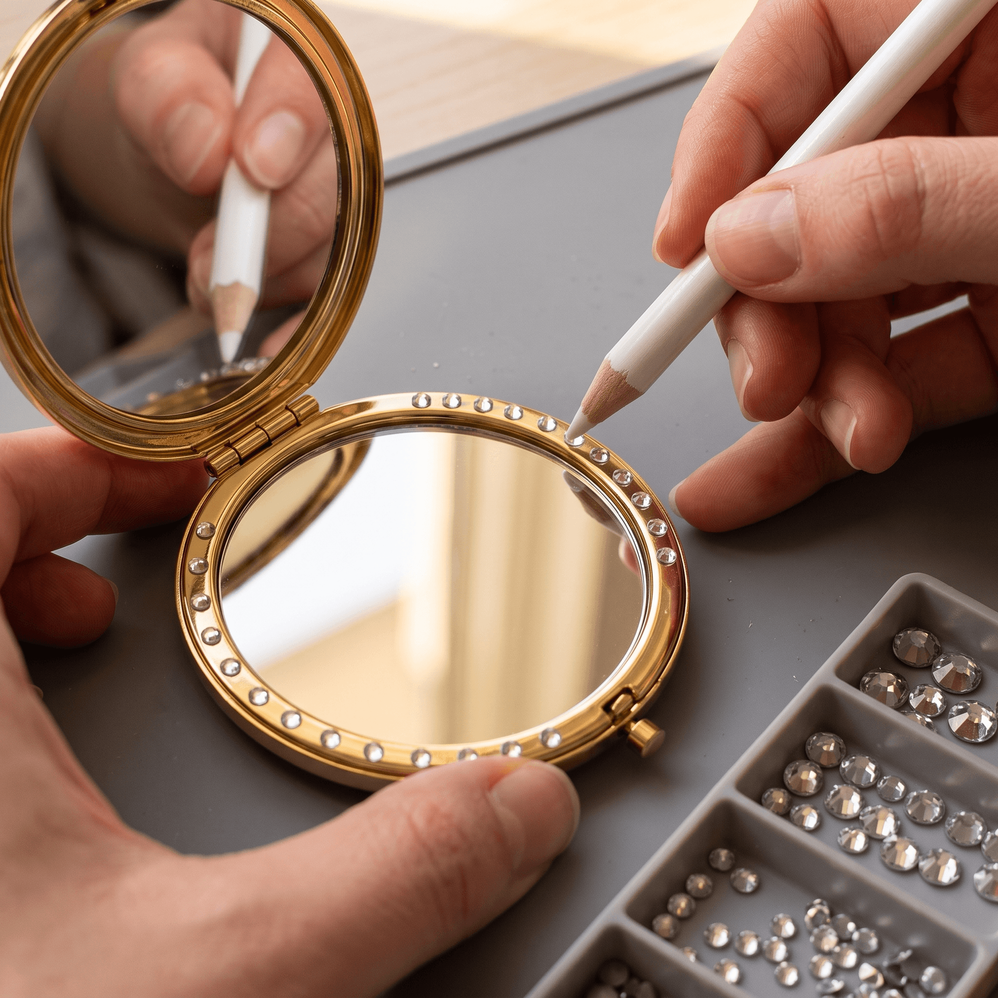 Artisan hands applying rhinestones to the edge of a gold compact mirror.